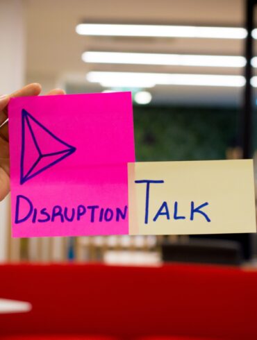 Image of a hand with two sticky notes, one in pink writing "disruption" and another in yellow writing "TALK".