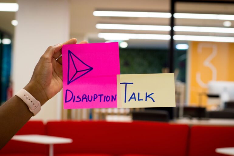 Image of a hand with two sticky notes, one in pink writing "disruption" and another in yellow writing "TALK".