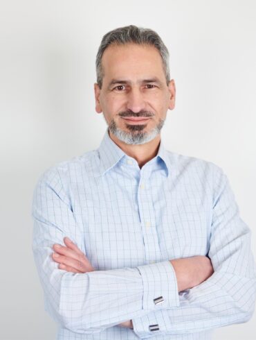 A professional business photo of Alex Mifsud as he poses wearing a light-blue chequered shirt with crossed arms