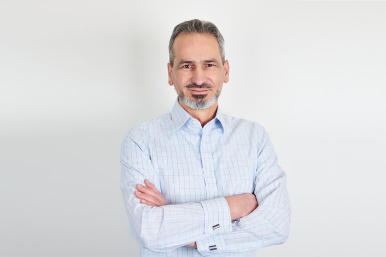 A professional business photo of Alex Mifsud as he poses wearing a light-blue chequered shirt with crossed arms