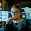 Photo of an intelligent looking woman wearing glasses as she stares into her computer screen with photos of a world map alongside charts that look related to economics.