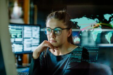 Photo of an intelligent looking woman wearing glasses as she stares into her computer screen with photos of a world map alongside charts that look related to economics.