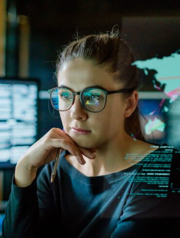 Photo of an intelligent looking woman wearing glasses as she stares into her computer screen with photos of a world map alongside charts that look related to economics.