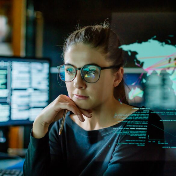 Photo of an intelligent looking woman wearing glasses as she stares into her computer screen with photos of a world map alongside charts that look related to economics.