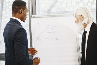 Photo of two men from diverse backgrounds in business attire making investment plans on a whiteboard with a marker