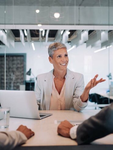 Photo of a woman wearing business attire in her office while providing a service to a couple whilst sat in front of a laptop
