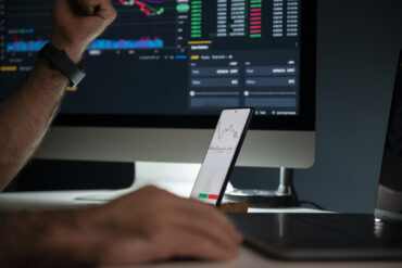 Photo of a man's hands as he trades on an exchange using both his desktop computer and smartphone at the same time.