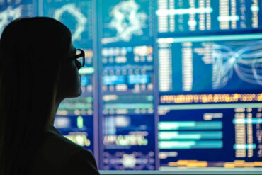 Photo of a woman in glasses looking at multiple screens with analytical data