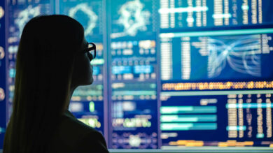 Photo of a woman in glasses looking at multiple screens with analytical data