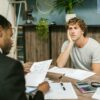 Photo of two-men sitting across eachother on a desk. The financier and client. The client looks depressed as the financier goes through his debt.