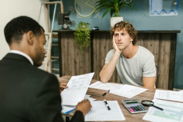 Photo of two-men sitting across eachother on a desk. The financier and client. The client looks depressed as the financier goes through his debt.