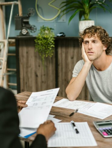 Photo of two-men sitting across eachother on a desk. The financier and client. The client looks depressed as the financier goes through his debt.
