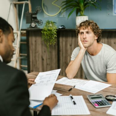 Photo of two-men sitting across eachother on a desk. The financier and client. The client looks depressed as the financier goes through his debt.