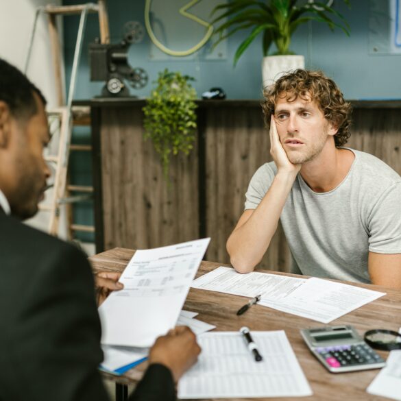Photo of two-men sitting across eachother on a desk. The financier and client. The client looks depressed as the financier goes through his debt.
