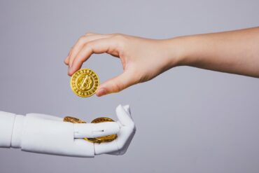 Photo of a human hand giving a crypto coin to a robotic hand, symbolising centralised finance working with decentralised finance