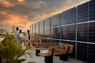 The terrace of a corporate building with solar panels on top designed in a modern fashion as three people walk around and point at them.