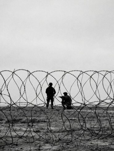 A black and white photo of soldiers taken from behind barbed wire, depicting a warzone and anti-terror campaigns.