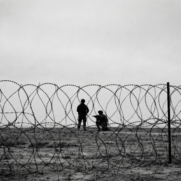 A black and white photo of soldiers taken from behind barbed wire, depicting a warzone and anti-terror campaigns.