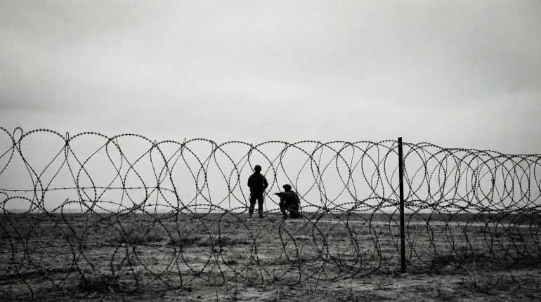 A black and white photo of soldiers taken from behind barbed wire, depicting a warzone and anti-terror campaigns.