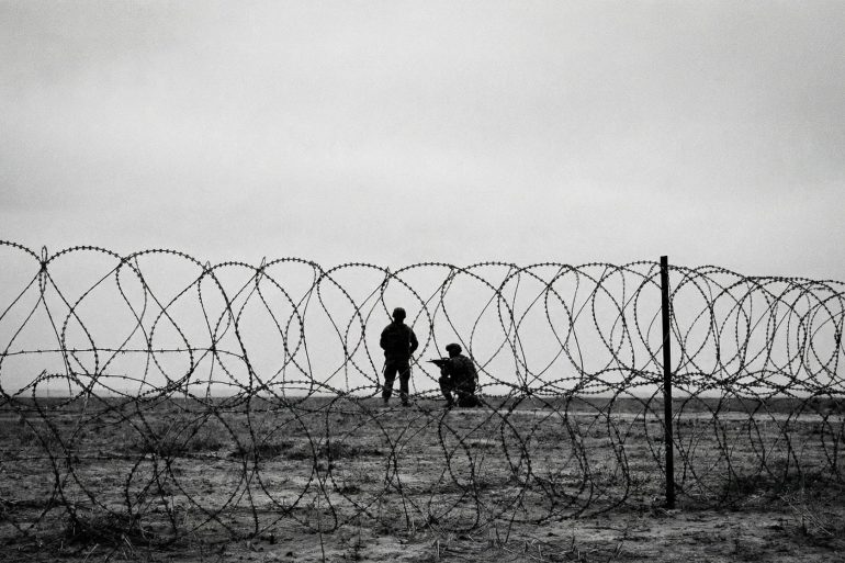 A black and white photo of soldiers taken from behind barbed wire, depicting a warzone and anti-terror campaigns.
